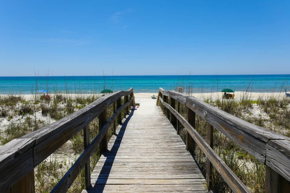 A wooden boardwalk crosses over sand dunes to the bright blue waters of Henderson State Park in Destin, FL.