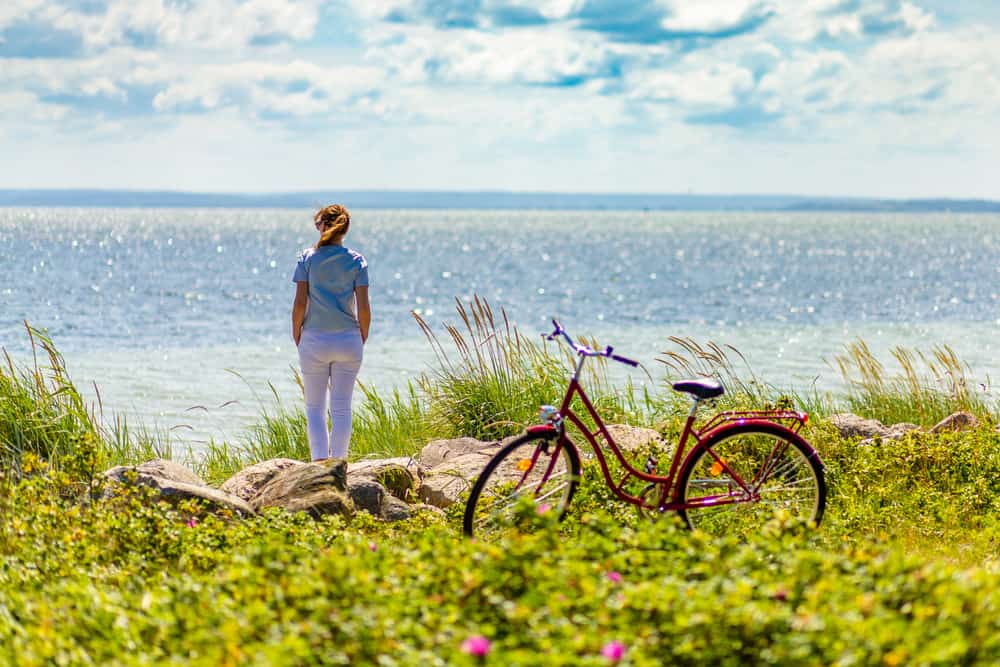 A woman looking out at the ocean near a bike that is parked in some grass near the beach. 