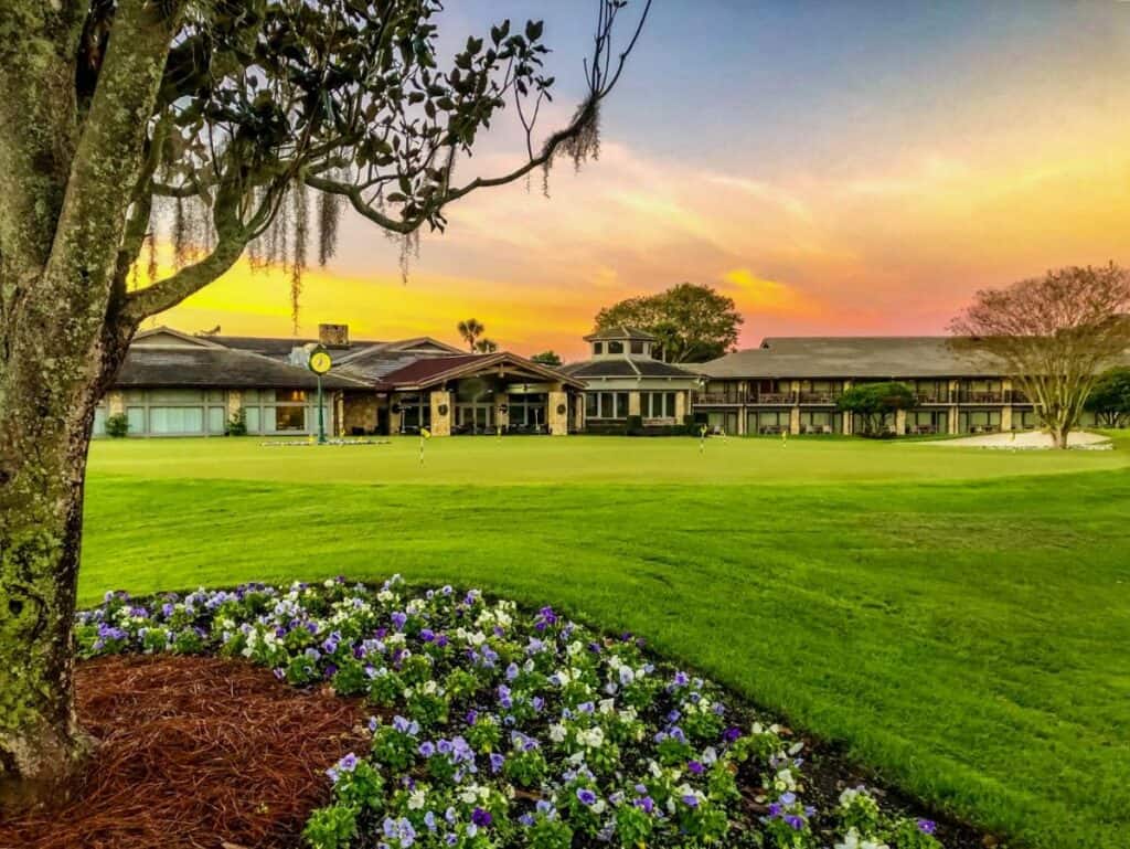 a shot of the arnold palmer bay hill club in the morning sun, with the greens of the golf course in the foreground, the well manicured lawn has a border garden of bright purple and yellow flowers. One of the best boutique hotels in Florida for golfers!