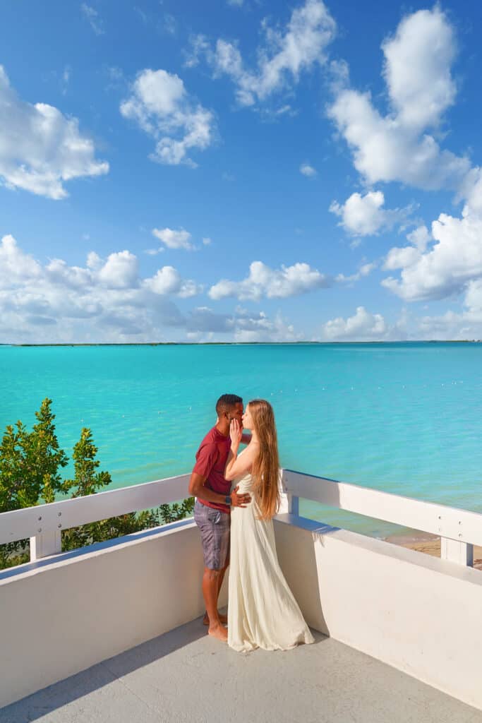 A shot of a couple embracing on the terrace of Baker's Cay, with the teal ocean in the background, beautiful blue sky in the background is sliced in half by the ocean's water, creating a stunning composition of blues
