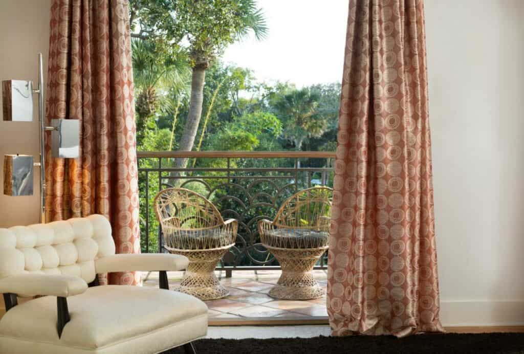 A daytime shot looking out onto the balcony of a room in the Black Dolphin Inn. The curtains are a 70's red and gold circle pattern, and the chairs outside on the balcony are vintage rattan. The view behind the balcony is lush vegetation