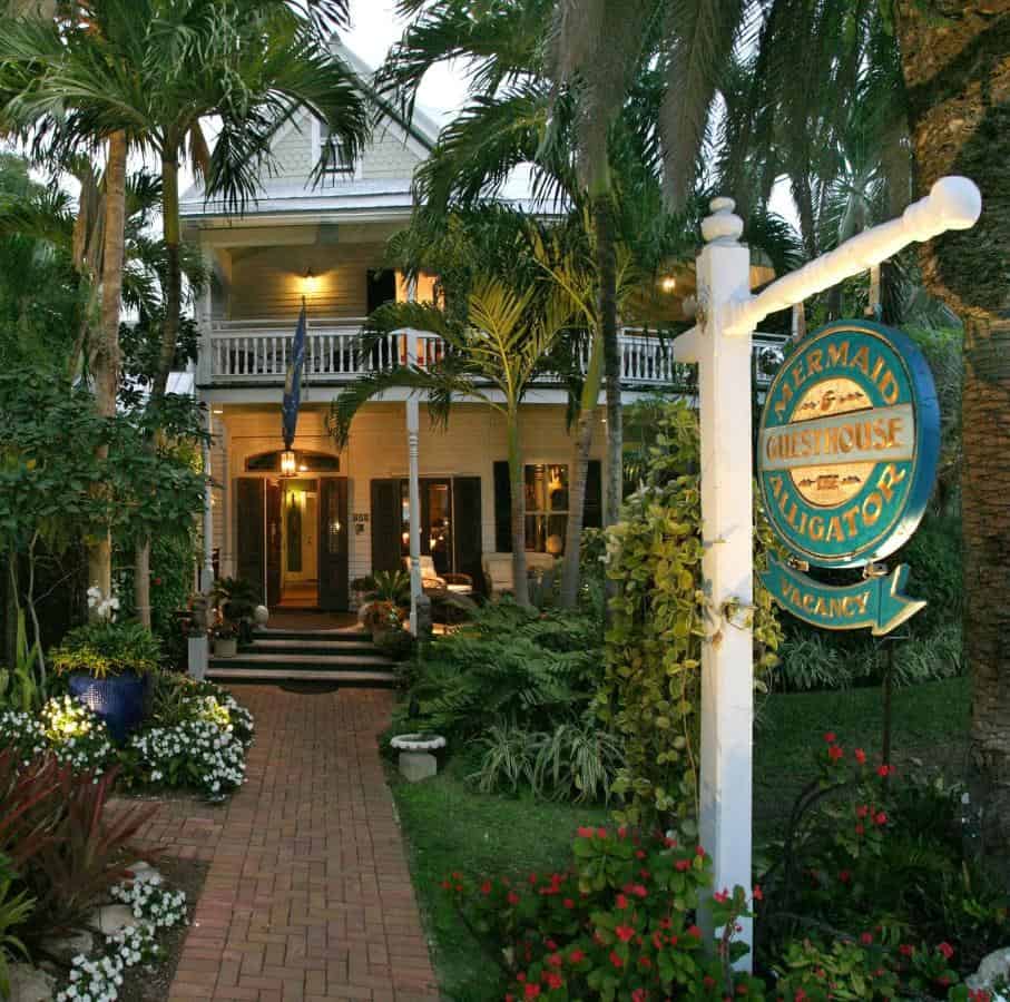 A dusky photo of the Mermaid and the alligator's entryway. The walkway is covered on all sides with greenery leading up to the entrance door in the background with a lit sign in the foreground of the hotel's logo. THe sign is beautiful and colorful with golden lettering
