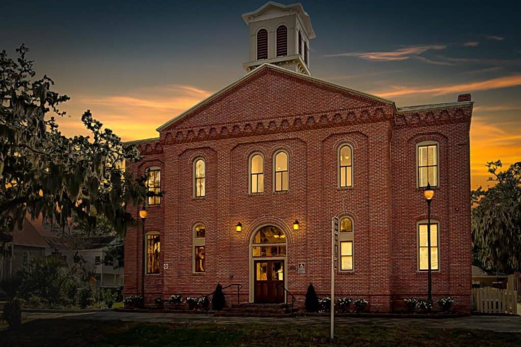 A night-time shot of the front of the schoolhouse inn, the building is made of red brick with a white belltower looming above, the sun is just setting, washing everything in warm sunset hues.
