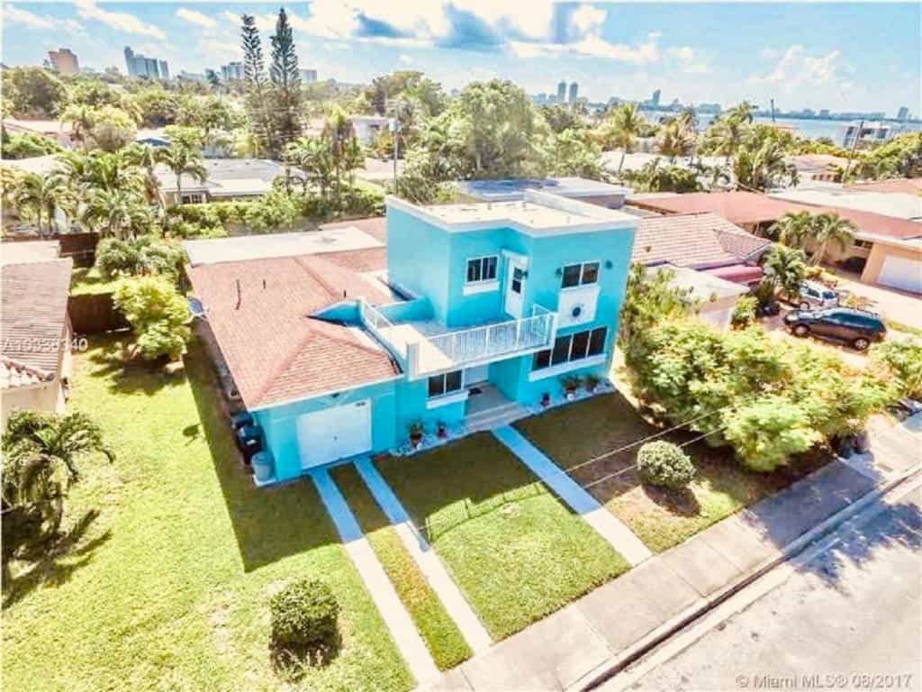 an aerial shot of a split level house that is decorated in all blue with a white trim and white balcony at the front. The house is surrounded by green grass and palm trees, with the ocean visible in the background 