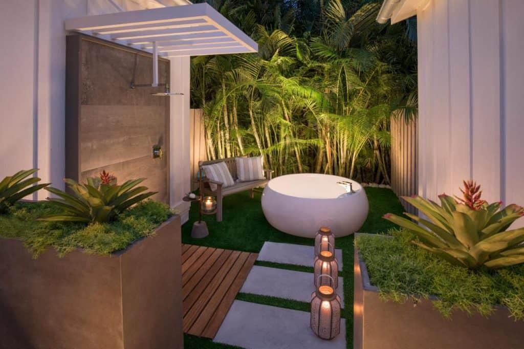 a beautiful photo of the soaking tub and outdoor shower in one of the private verandas attached to the bungalows. There is lots of lush greenery, warm toned lighting, and the bathtub itself looks like a large porcelain egg. 