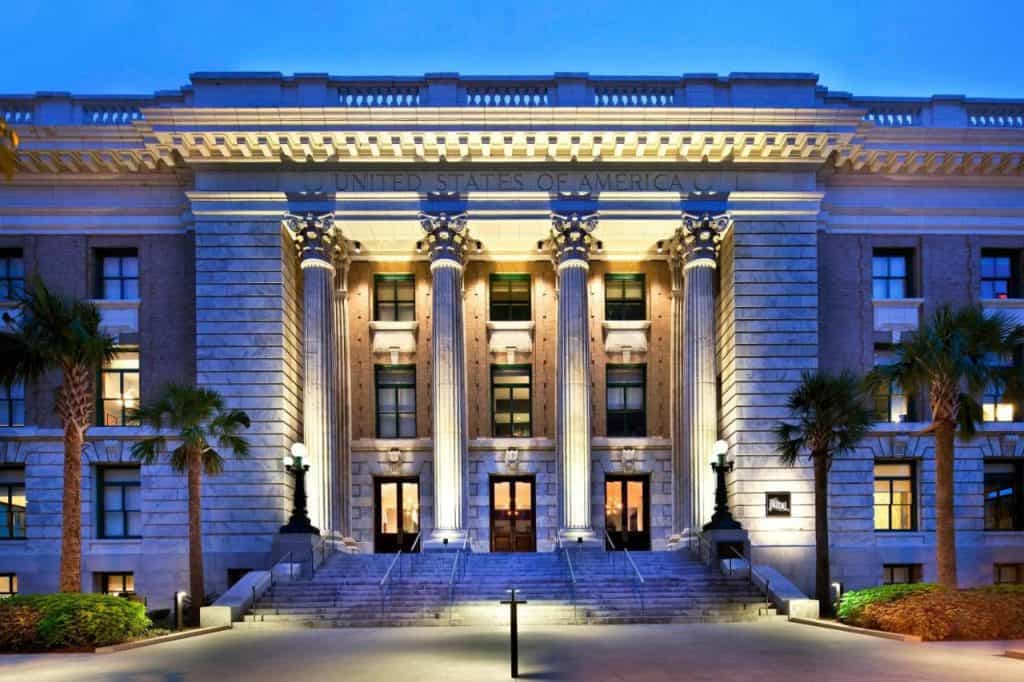 A twilight shot of the le Meridien Hotel's exterior which is that of a courthouse. The four pillars in the center house three doors which lead into the lobby. There are palm trees in the foreground, creating a lush vibe