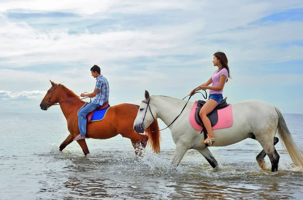 A couple riding a pair of horses through the water on the beach, one of the best things to do in Amelia Island. 