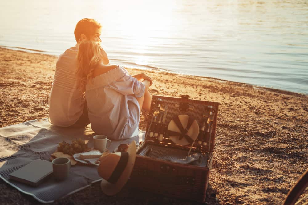 A couple sitting next to each other on a picnic blanket on the beach with a picnic basket as the sun is setting. 