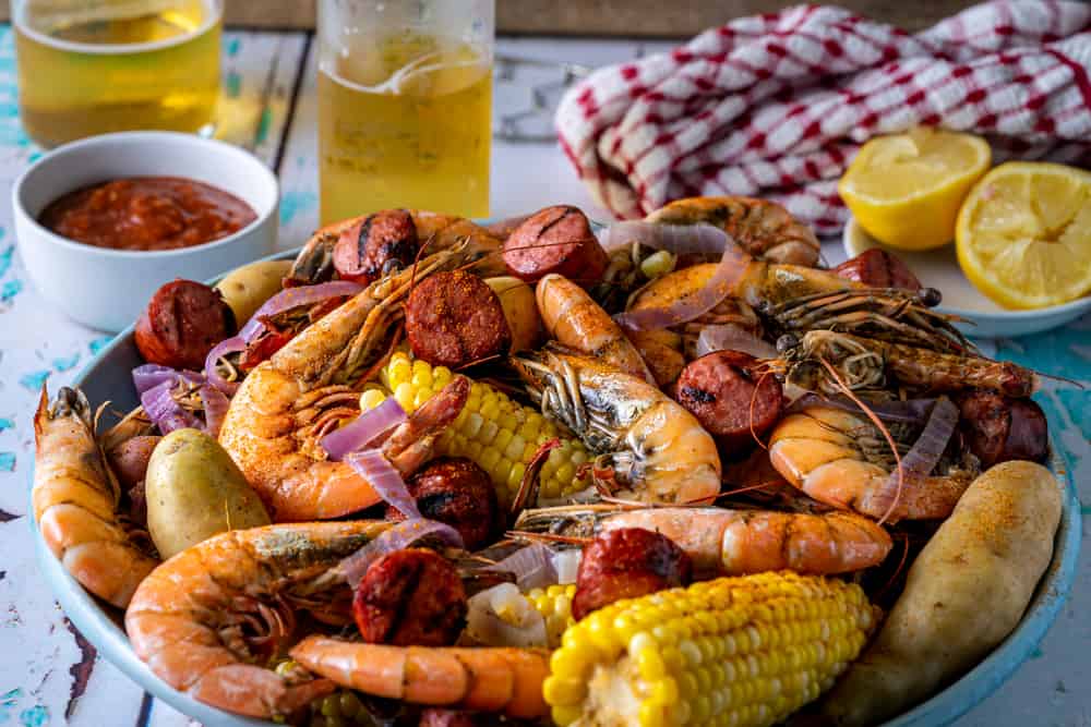 A platter of shrimp, corn, and sausage sits on a rustic table, with sides of lemon, cocktail sauce, and a glass of beer.