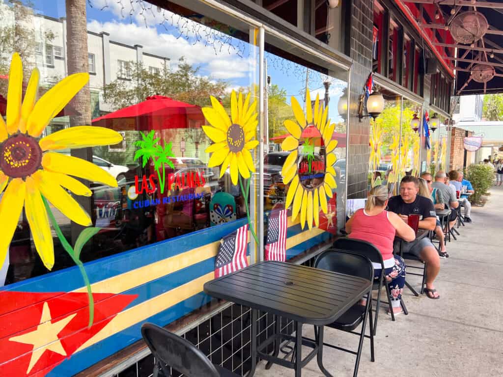 The outside of Las Palmas Cuban Restaurant one of the restaurnats in Mount Dora. People are sat on the sidewalk and there are colorful painting on the windows. 