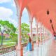 woman standing in blue dress leaning against a pink column and looking out over a courtyard with green trees