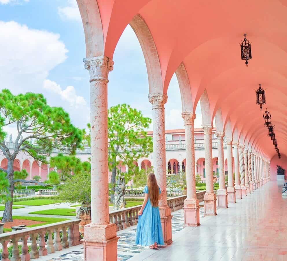 woman standing in blue dress leaning against a pink column and looking out over a courtyard with green trees