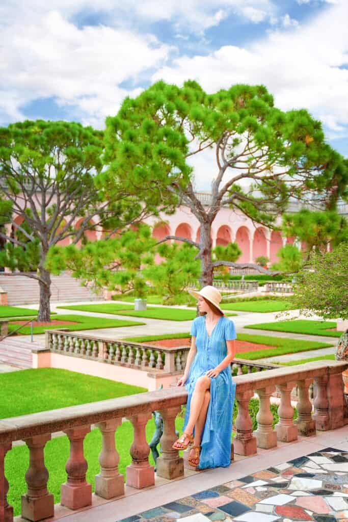 Victoria perches herself on the edge of a walkway to look out at the center gardens of the Ringling Museum. She is wearing a blue dress and sunhat.