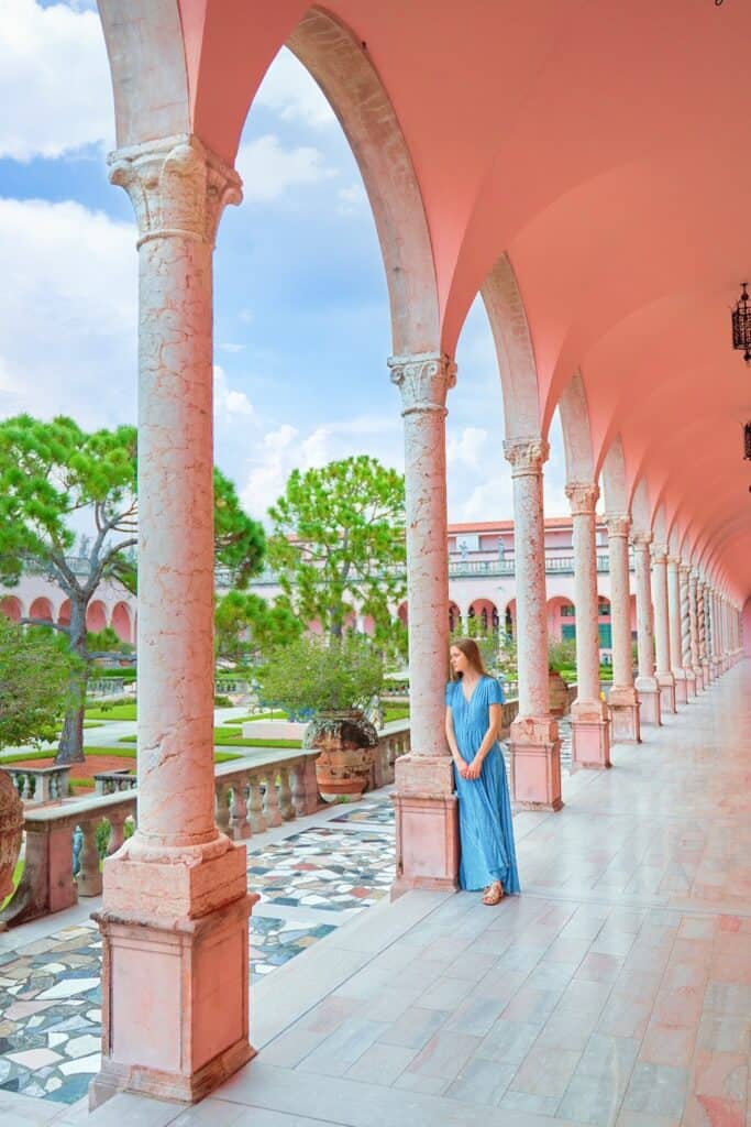 Victoria, in a blue dress, leans against a stone pillar and gazes out at the gardens at the Ringling Museum. 