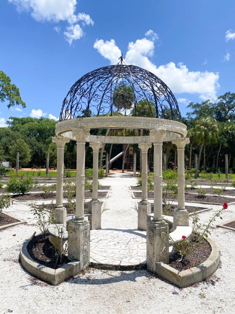 A gazebo at the Rose Garden at the Ringling Museum is made of stone and iron and is surrounded by pathways and flowers. 