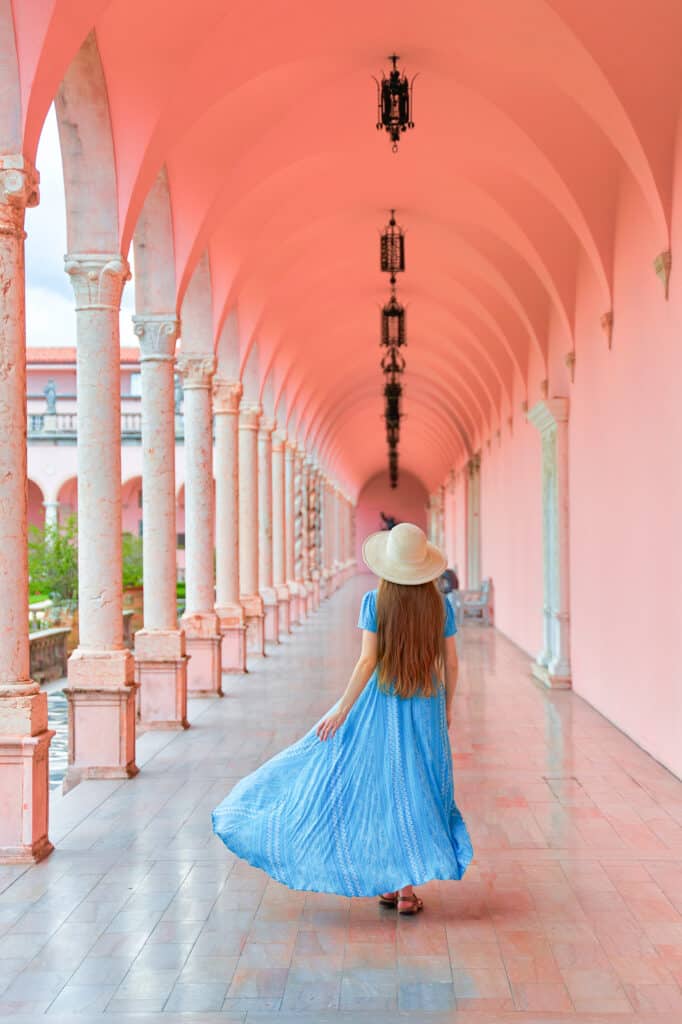 A picture of a woman in a light blue dress standing in a long marble walkway with light pink walls at the ringling museum, one of the best things to do in Florida!