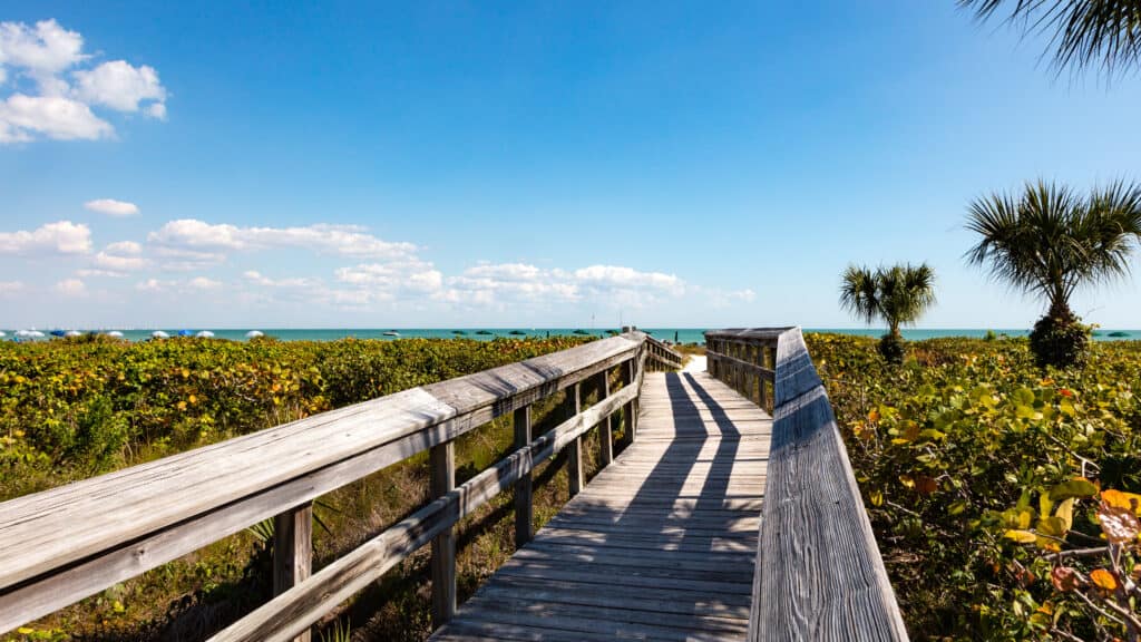 A daylight photo of the path leading down to the beach at Sanibel Island, the path is surrounded by green foliage on either side 