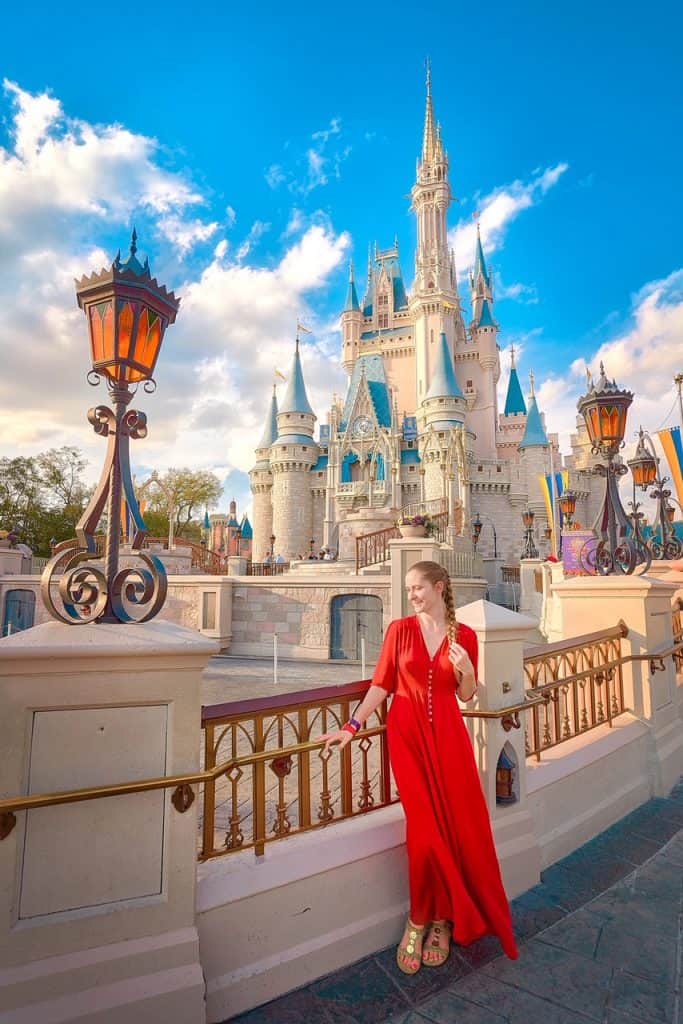 A woman in a long red dress leans against a railing in front of a fairytale castle, with spires and blue roofs, bathed in warm sunlight.