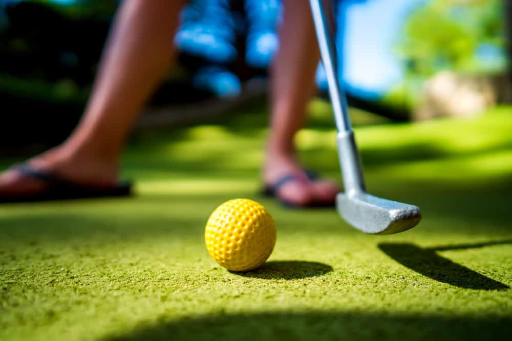 A yellow mini golf ball sits on a green turf course with feet wearing flip flops in the background. Playing mini golf is one of the best activities to do in Clearwater Beach.