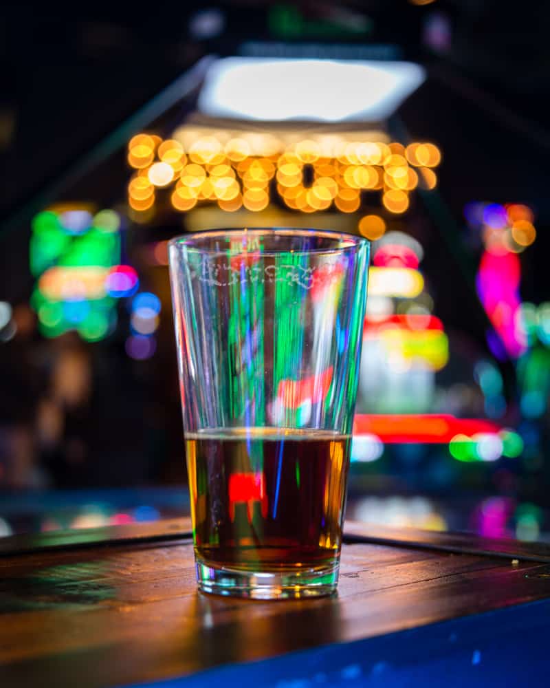 A glass of beer sits on a table, half full, with arcade lights blinking in neon colors behind it at one of the bars in Gainesville that is also an arcade. 