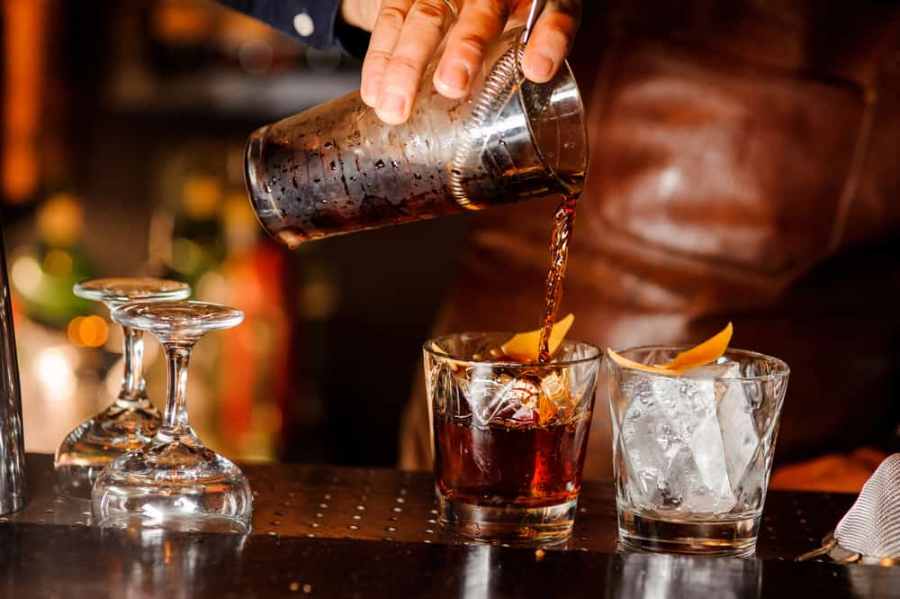 At one of the handcrafted cocktail bars in Gainesville, a bartender preps drinks by pouring a mixture into two glasses that have ice and orange slices in them. 