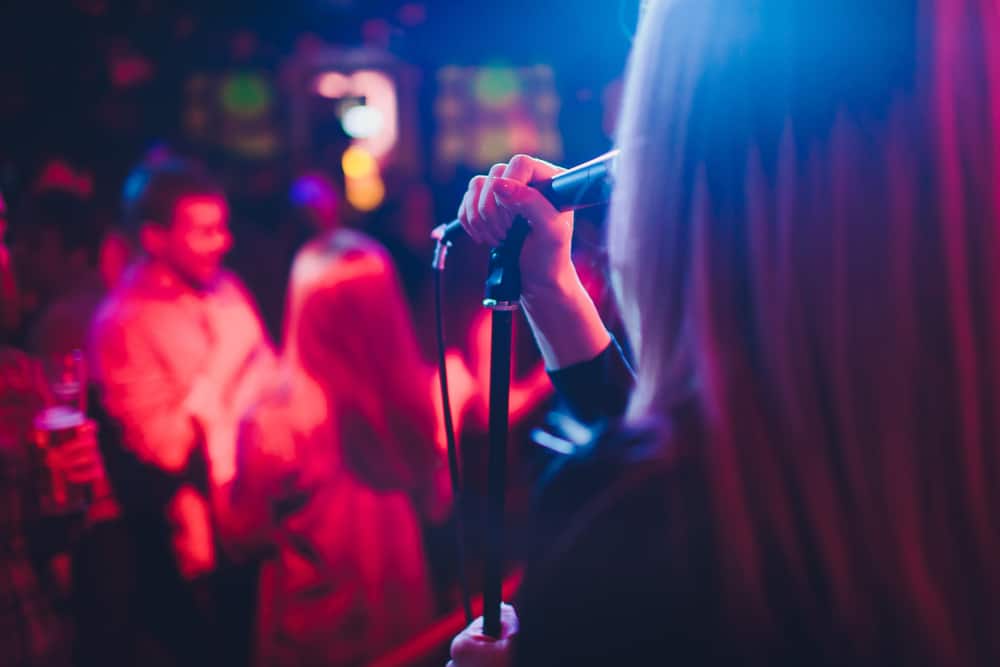 At one of the 80s themed bars in Gainesville, a young girl stands with her back to the camera as she sings karaoke on stage. You can see people in the crowd dancing and nursing drinks as she sings. 