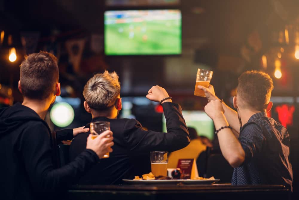 A trio of guys sit at a bar table and watch a flat screen tv at one of the many sports bars in Gainesville. They toast with their beers and cheer. 
