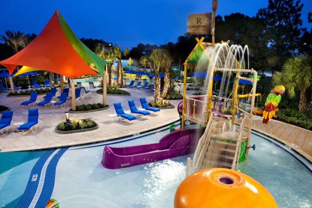 A nighttime shot of the pool area for the Renaissance Orlando, there are blue pool chairs and colorful playground pieces in the shallow splashing pool