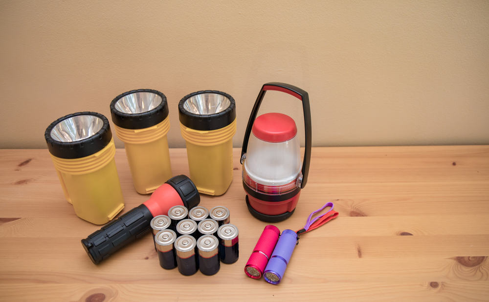 A series of flashlights and LED lanterns sit on a wooden table, alongside a variety of batteries. Be prepared with your hurricane preparedness list for power outages and ways to combat them. 