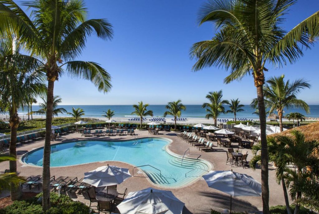 A picture of the irregularly shaped pool at Lido Key Beach resort, the blue and white umbrellas blend with the blue and white of the sandy beach in the background, one of the best hotels in Florida on The beach