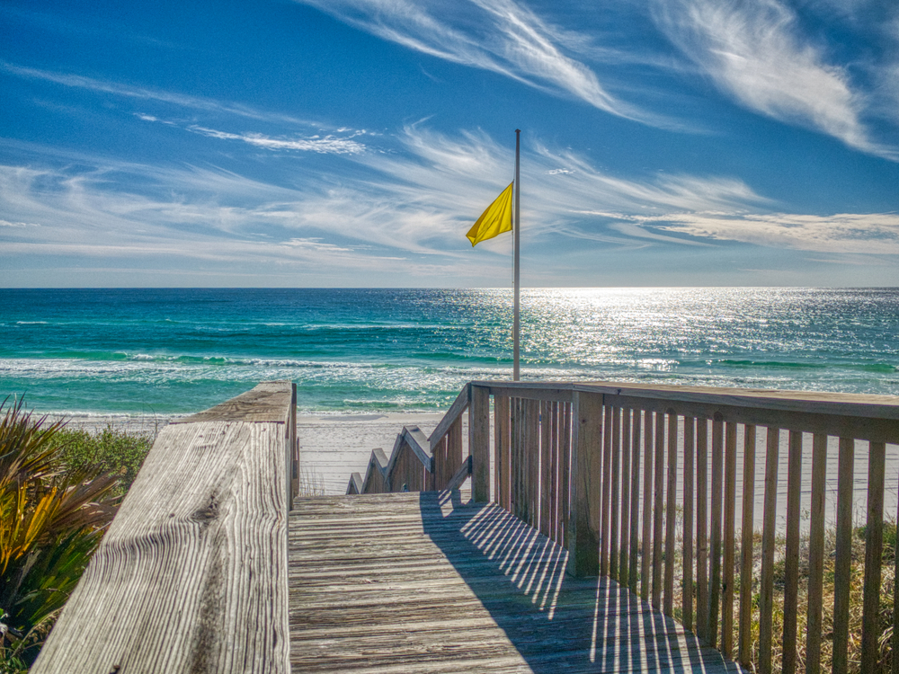 A yellow flag stands next to the wooden stairs that lead down to Blue Mountain Beach on a sunny day in Florida.