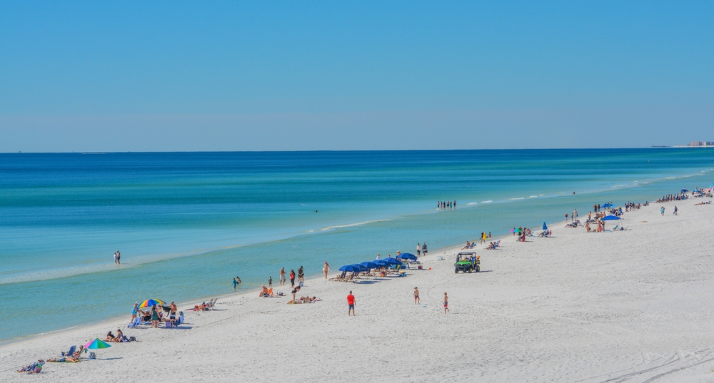 Several people stand in the calm blue waters, with many other people reclining on the sad at Miramar Beach, one of the best beaches near Destin.