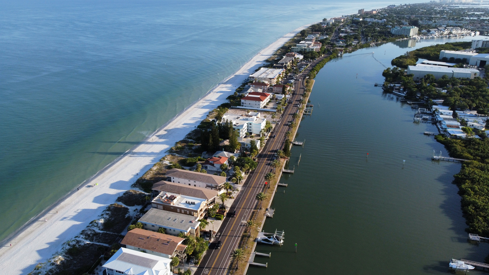 An aerial view of Indian Rocks Beach on a sunny day, a thin strip of beach land lined with buildings along blue water.