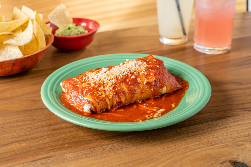 A burrito covered in cheese and red sauce sits on a teal plate, with chips ad guacamole in the background, like those served at VIP, one of the best Indian Rocks Beach restaurants for Mexican.