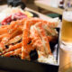 A container full of crab legs sits next to a glass of beer on a wooden table.