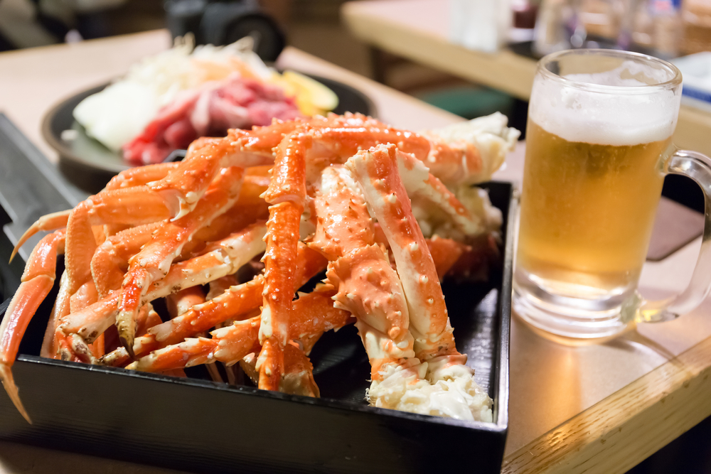 A square plate full of crab legs sits next to a glass of beer on a wooden table, like those you can order at Crabby Bill's, one of the most popular Indian Rocks Beach restaurants.