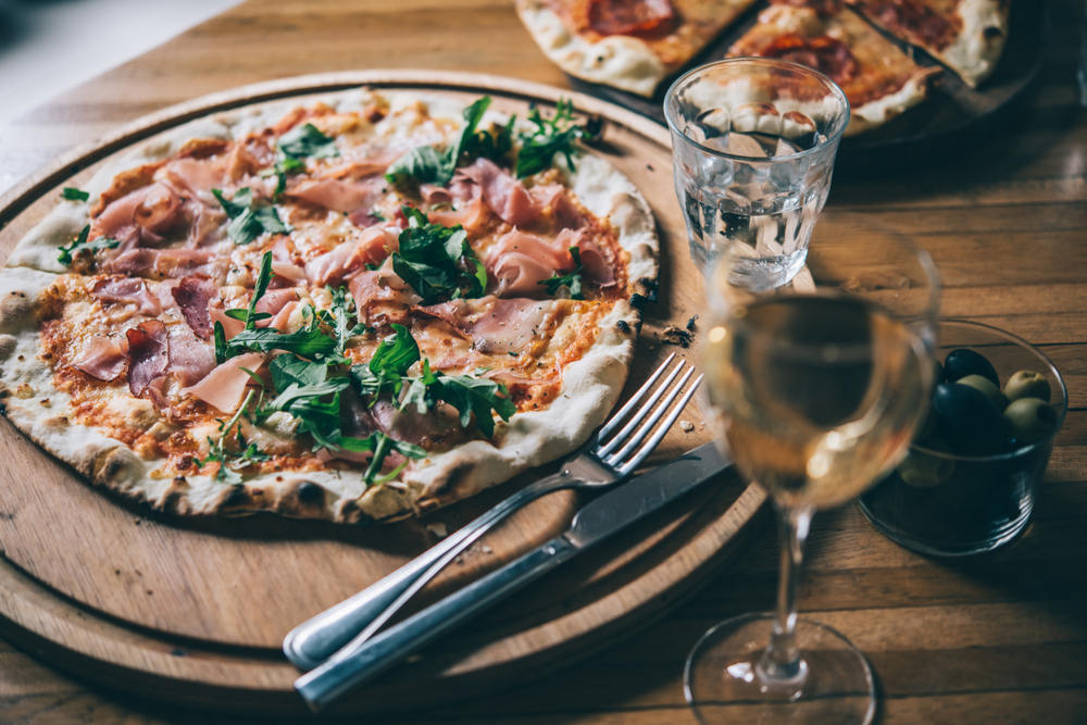 A thin crust pizza with arugela annd prosciutto sits on a wooden platter, next to a glass of white wine, similar to a scene at Slyce Indian Rocks Beach, one of the most popular Indian Rocks Beach restaurants.