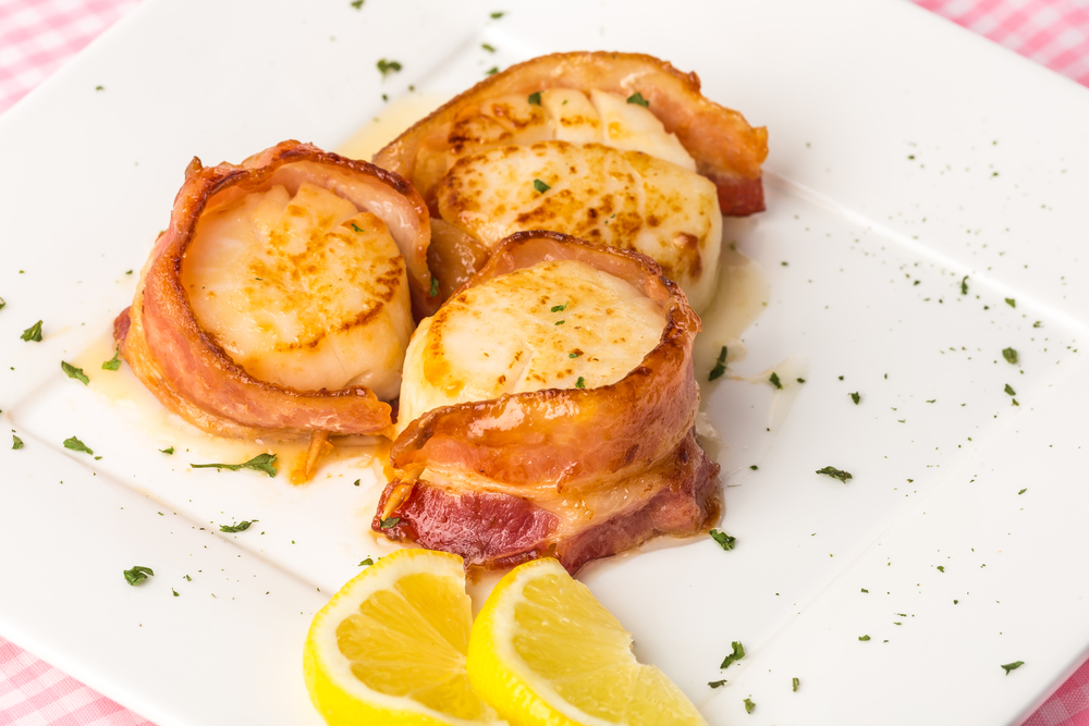 Three bacon-wrapped scallops sit on a white plate with two lemon wedges.