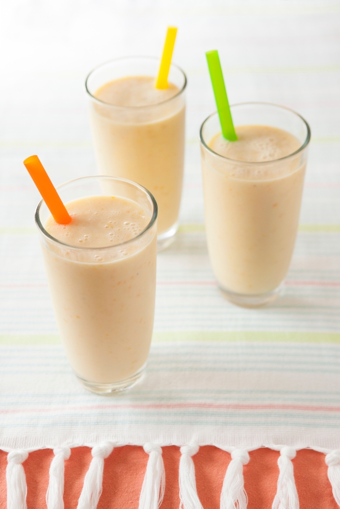 Three orange creamsicle smoothies with different colored straws stand in glasses on a light-colored tablecloth.