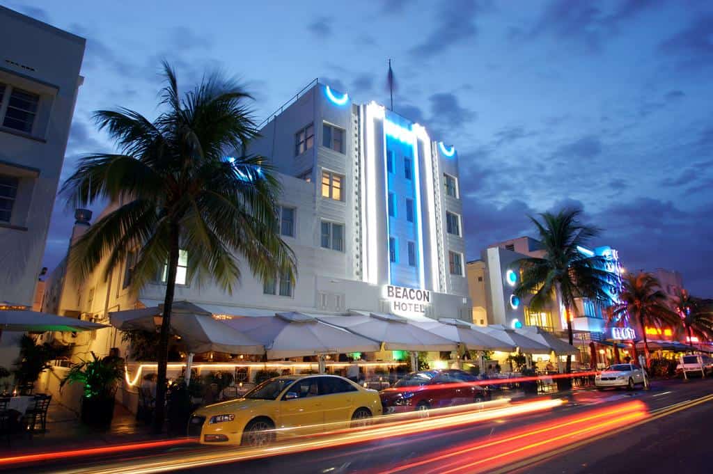 Cars pass by the Beacon Hotel in Miami at night, an art deco hotel with umbrellas and palm trees outside.