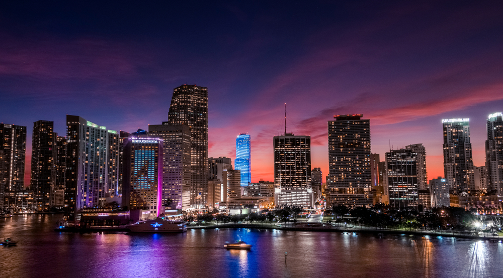 The Miami skyline at sunset, with a single boat in the bay where the lights are reflected in the water.