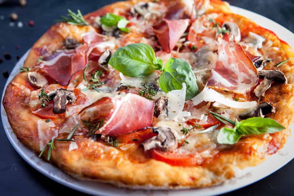 A closeup shot of a pizza, with mushrooms, prosciutto, cheese, and a garnish of basil.