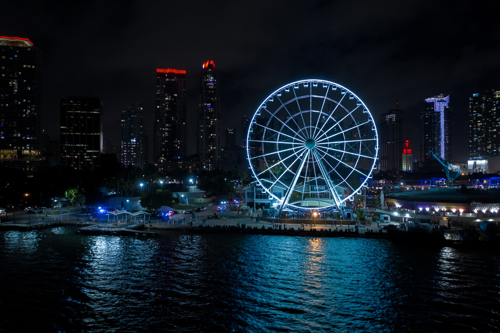 The illuminated Skyviews Miami Observation, one of the best things to do in Miami at night, rotates slowly standing near the water with skyscrapers in the background.