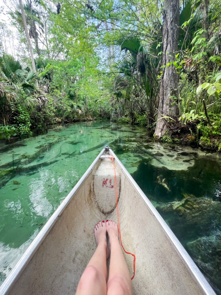 A woman's feet sit in the tip of a canoe as it heads down the river: you can see the sandy bottom of the river through the clear water as the boat moves. 