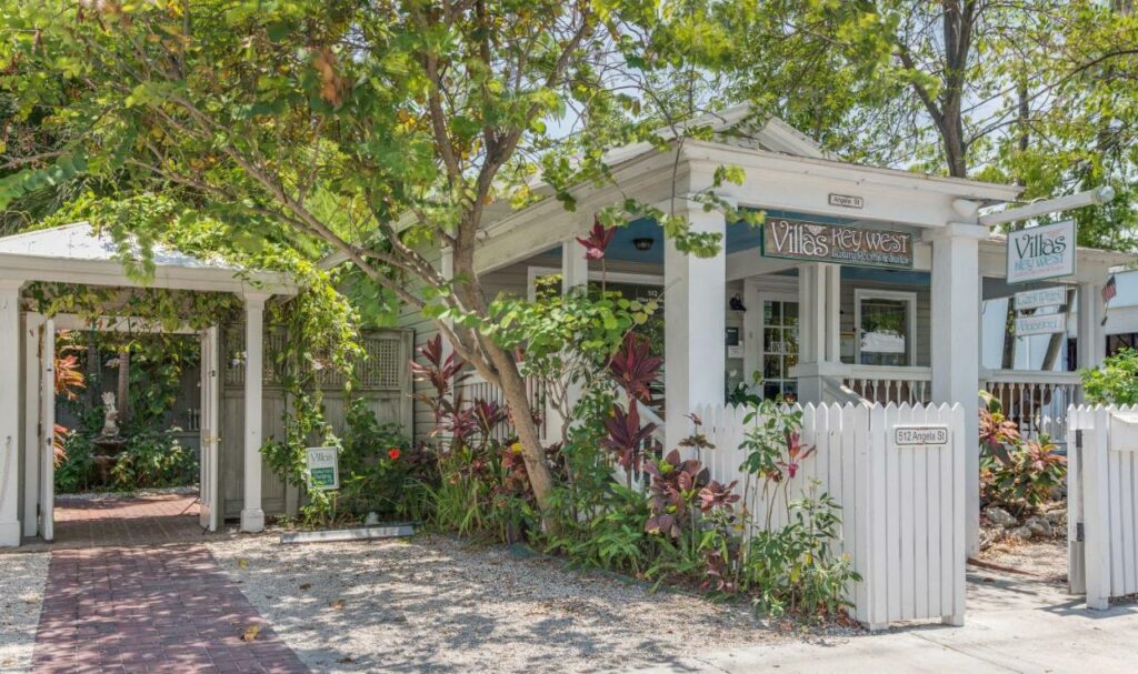 A shot of the plant-covered exterior of the Key West Villas, there is a sign with the name and the property is covered by a white picket fence