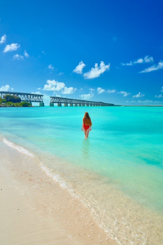A woman with long hair in a red bathing suit, looks at an old elevated railway while she stands knee-deep in bright blue water at Bahia Honda State Park, one of the best beaches in the Florida Keys.