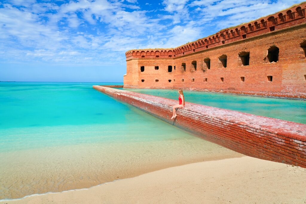 A woman in a red swimsuit sits on a brick wall that surrounds the brick walls of a fort at Dry Tortugas National Park, which has some of the clearest water of the beaches in the Florida Keys.