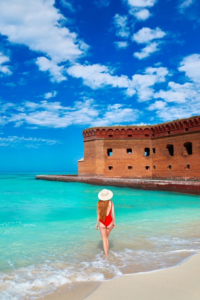 A woman with long hair wears a sunhat and red swimsuit as she walks in the surf, looking at the blue water and old brick fort at Dry Tortugas National Park in FL.