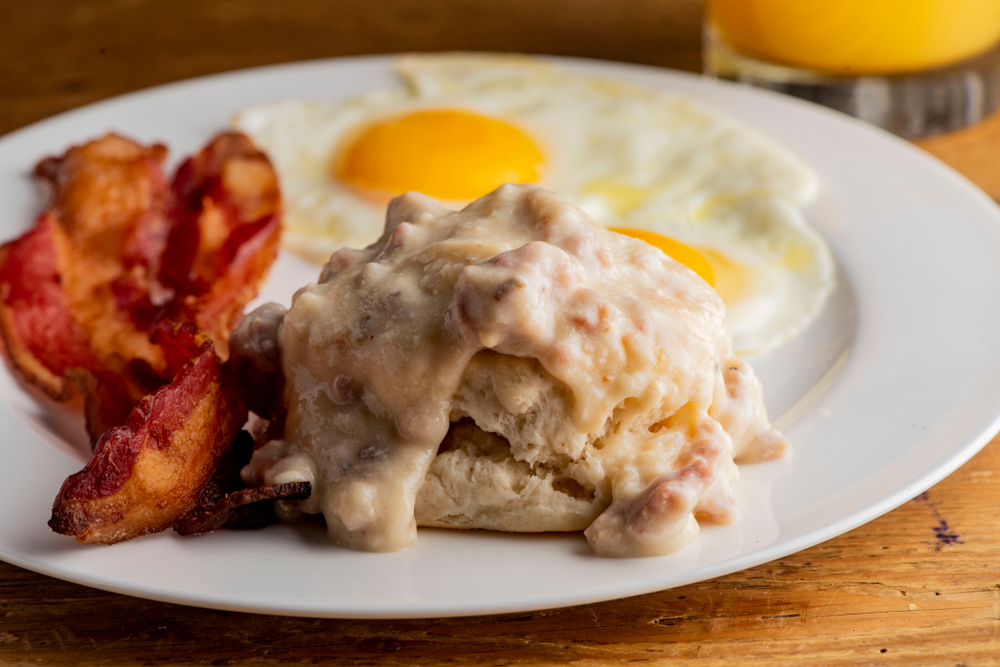 A biscuit with gravy, bacon, and two eggs sunny-side-up sit on a white plate. 