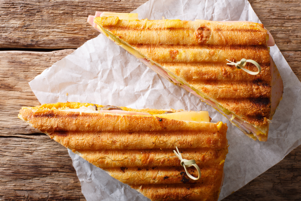 Two triangular Cuban sandwiches, with toothpicks holding them together, sit on a piece of paper on a wooden background.